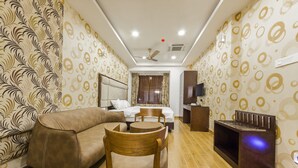 Une chambre d’hôtel avec un canapé, une chaise en bois, un lit avec des rideaux, une télévision et un ventilateur de plafond.