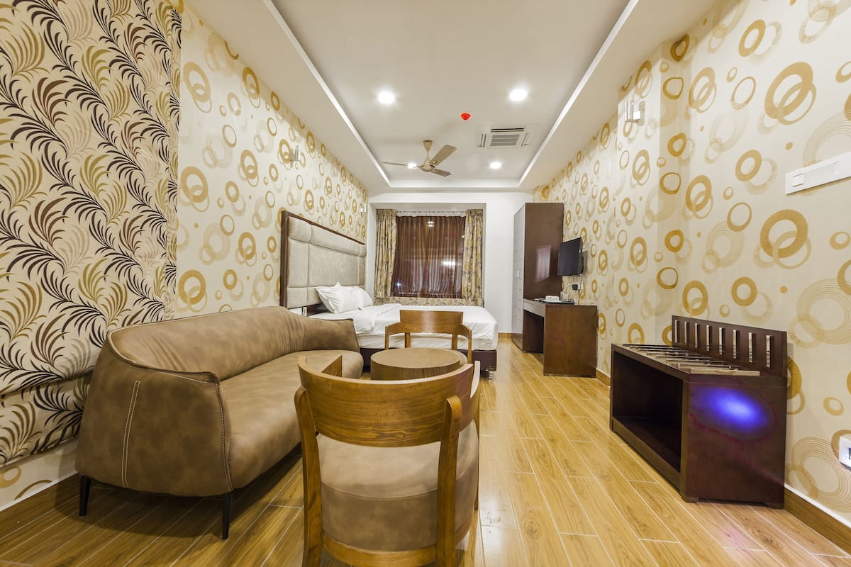 A hotel room with a sofa, a wooden chair, a bed with curtains, a TV, and a ceiling fan.