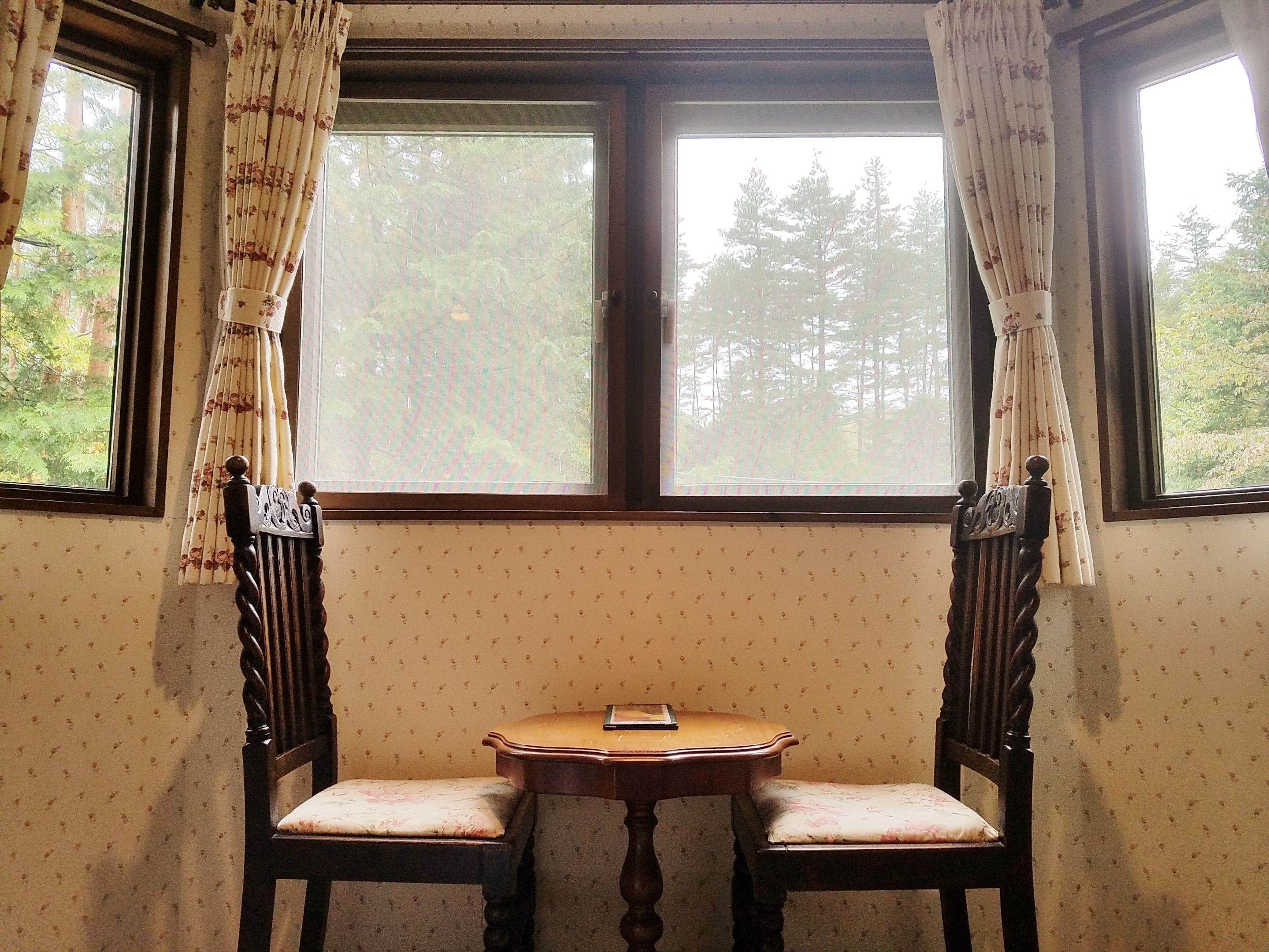 A room with two wooden chairs and a small table, a window with curtains, and patterned wallpaper.