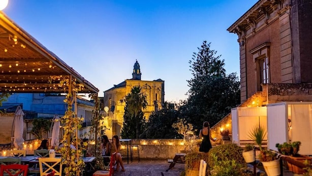 An outdoor patio area with tables and chairs, a building with classical architecture in the background, and string lights. - Dimora San Giuseppe (Lecce)