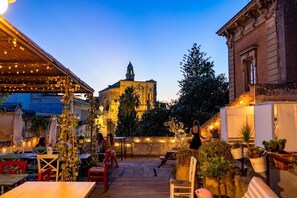 An outdoor patio area with tables and chairs, a building with classical architecture in the background, and string lights. - Dimora San Giuseppe (Lecce)