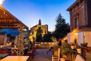 Un patio al aire libre con mesas y sillas, un edificio de arquitectura clásica al fondo y luces colgantes.