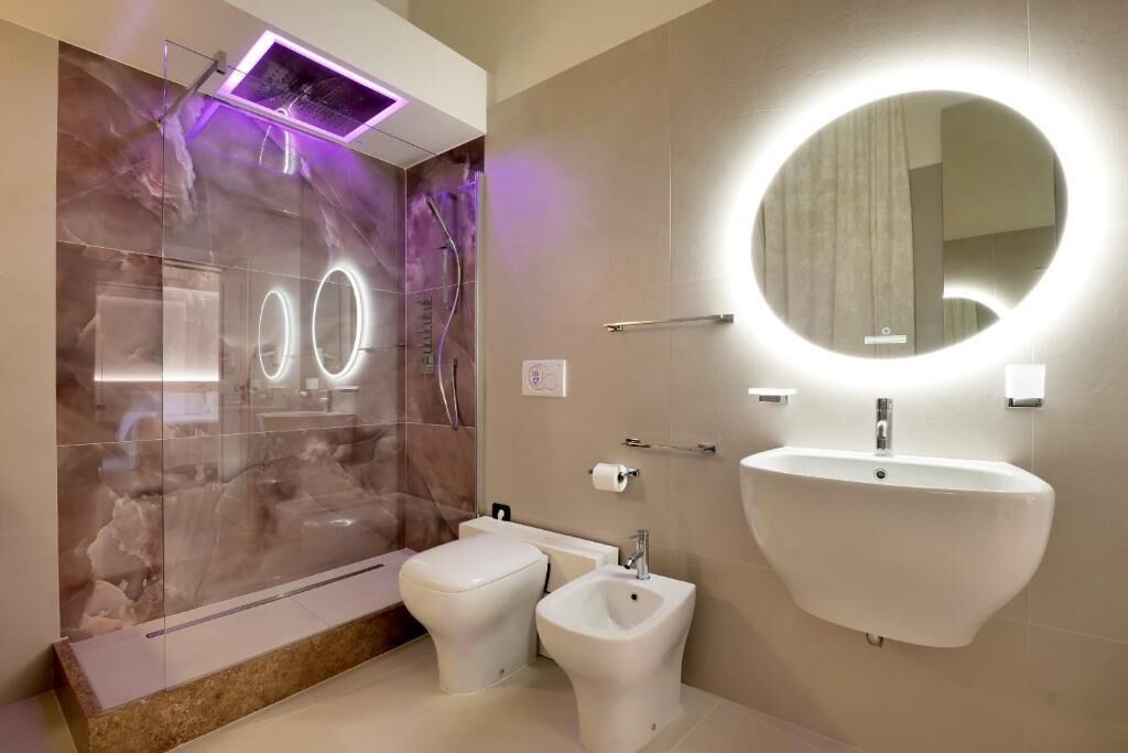 A modern bathroom with a glass shower enclosure, a pedestal bidet, a wall-mounted sink, and a round mirror with integrated lighting.