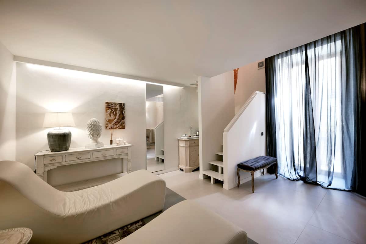 A modern living room with a sofa, a bench, a console table with decorative items, a staircase, and sheer curtains.
