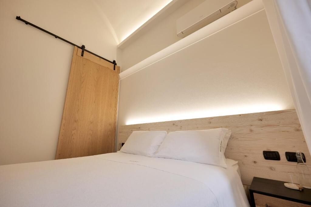 Une chambre d’hôtel moderne avec une tête de lit en bois, un lit recouvert de draps blancs, une table de chevet avec un verre et un luminaire fixé au mur.