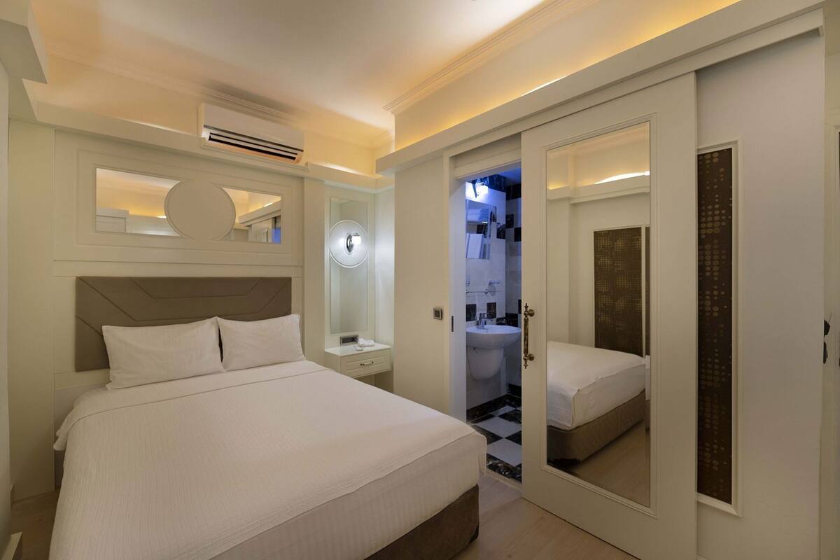 A hotel room with a large bed, a bedside table, a mirror, a wardrobe, and a bathroom visible through an open door.