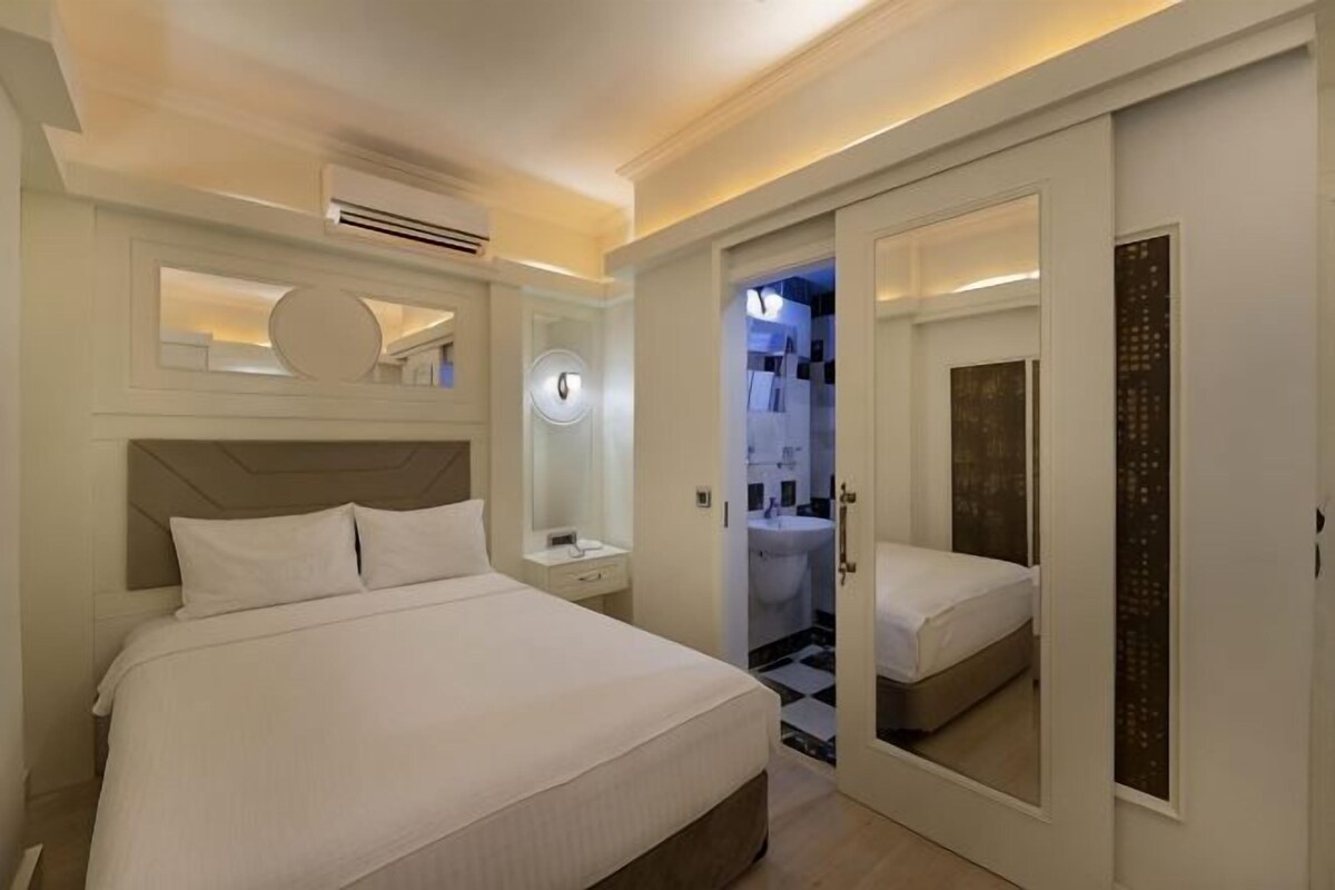 A bedroom with a bed, bedside table, mirror, and a bathroom with a sink and toilet.