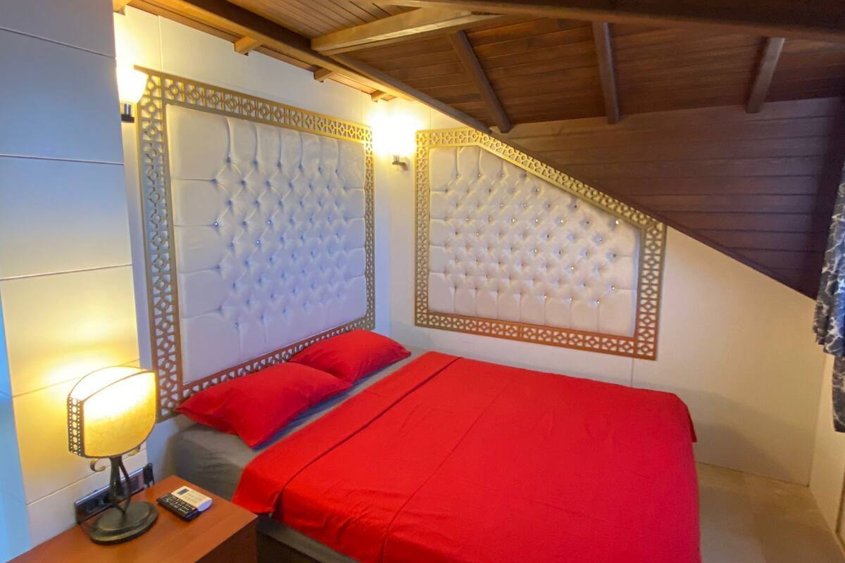 A bedroom with a wooden ceiling, a bed with a red bedspread, a nightstand with a lamp, and a decorative headboard.