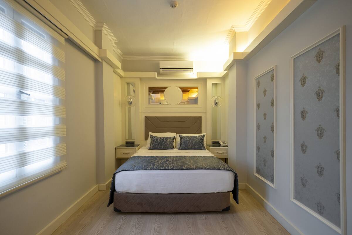A bedroom with a large bed, bedside tables, an air conditioning unit, and patterned wallpaper.