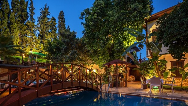Une piscine avec un pont en bois, entourée d’une végétation luxuriante et bordée d’un bâtiment doté d’un toit en tuiles.