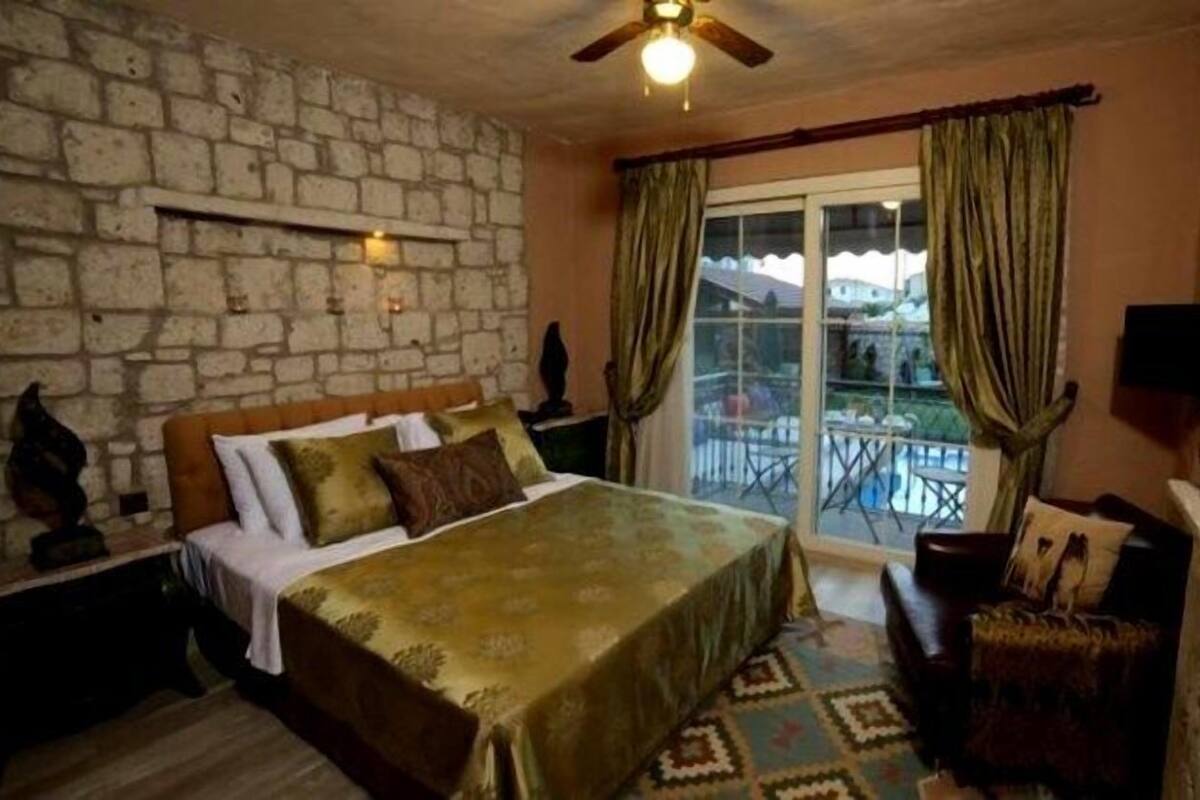 A bedroom with a stone wall, a bed with a green bedspread, a chair, a small table, and a balcony with a view.