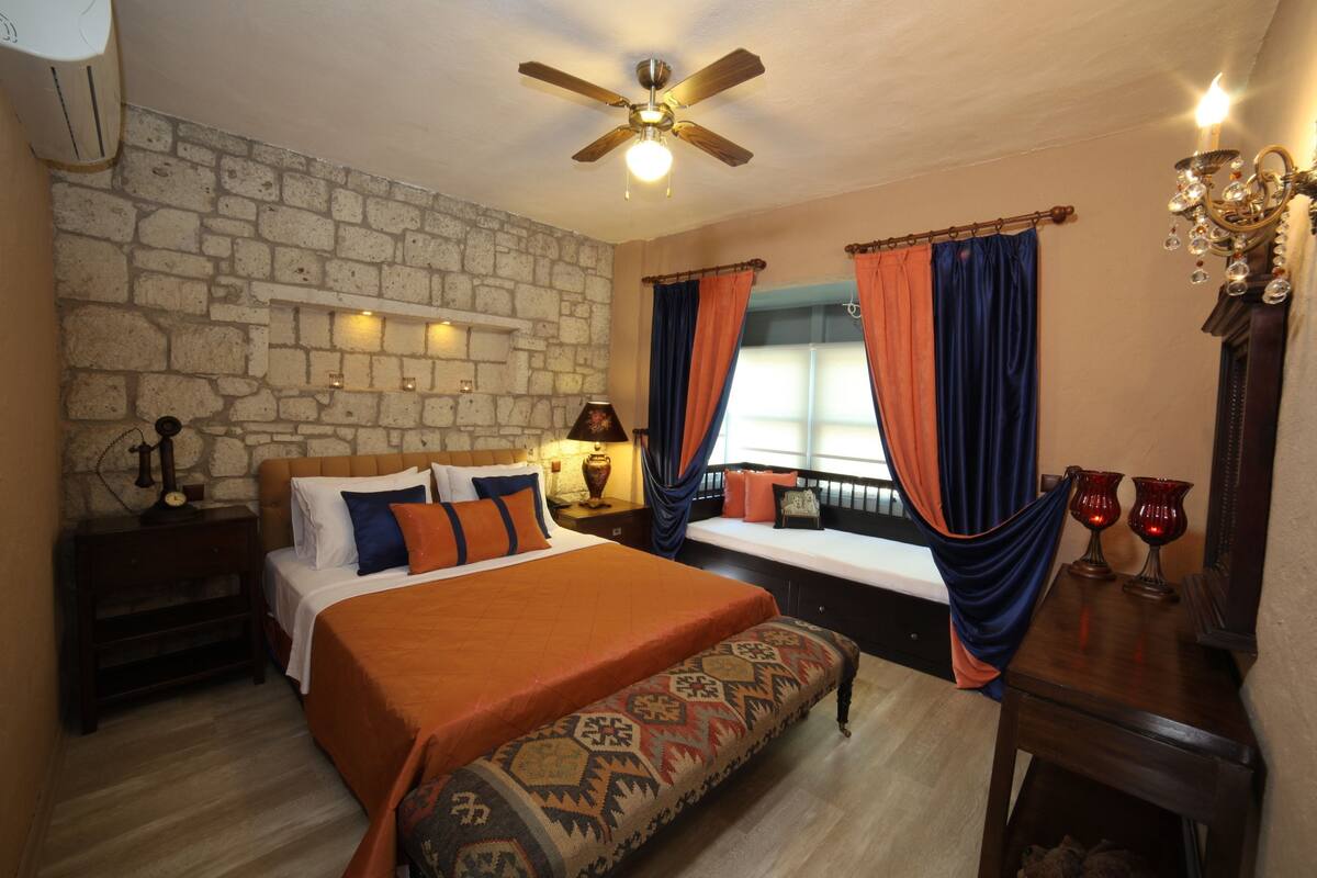 A bedroom with a stone wall, a bed with an orange bedspread, a bench, a nightstand with a lamp, and a window with blue and orange curtains.