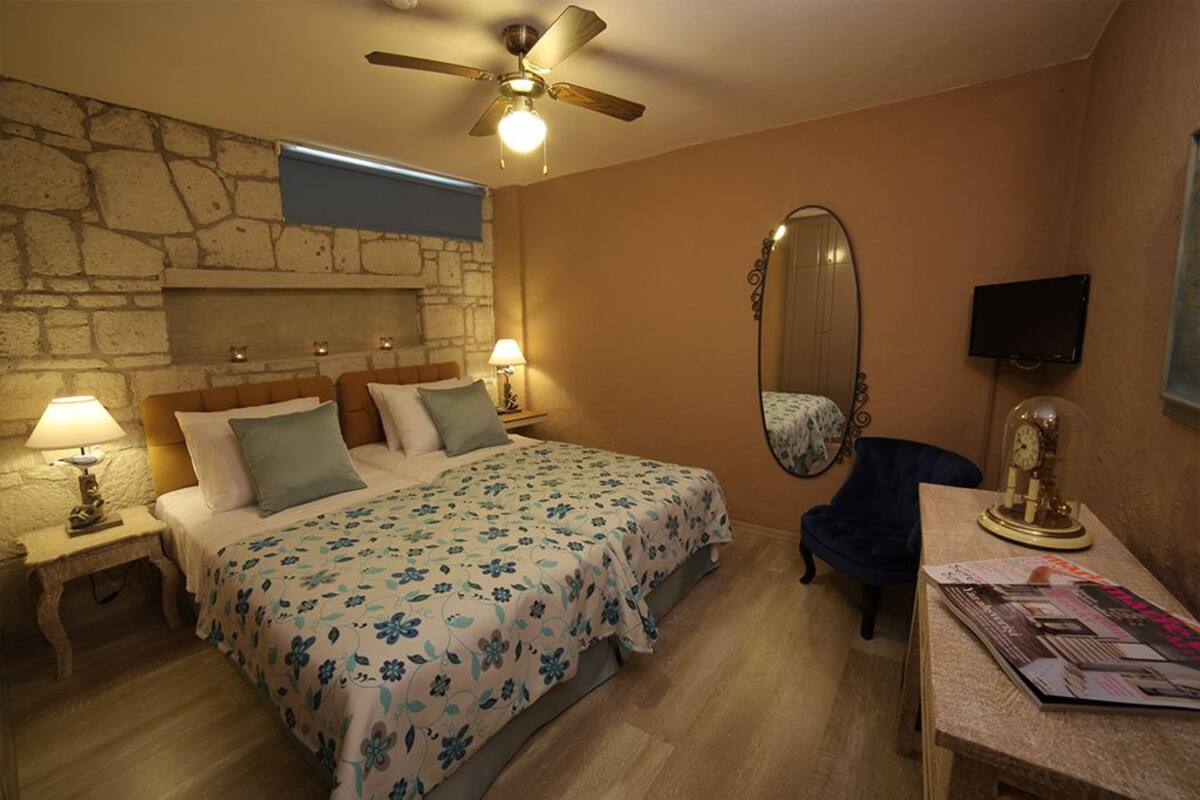 A bedroom with a stone wall, a bed with floral bedding, a ceiling fan, a mirror, a TV, and a desk with a clock.