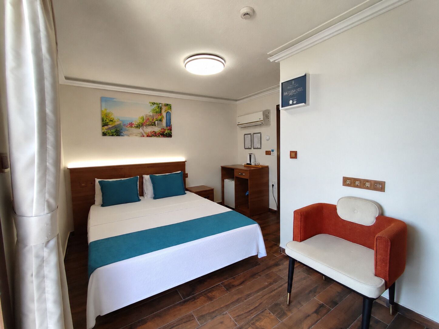 Una habitación de hotel con cama, escritorio, silla, equipo de aire acondicionado y un cuadro en la pared.