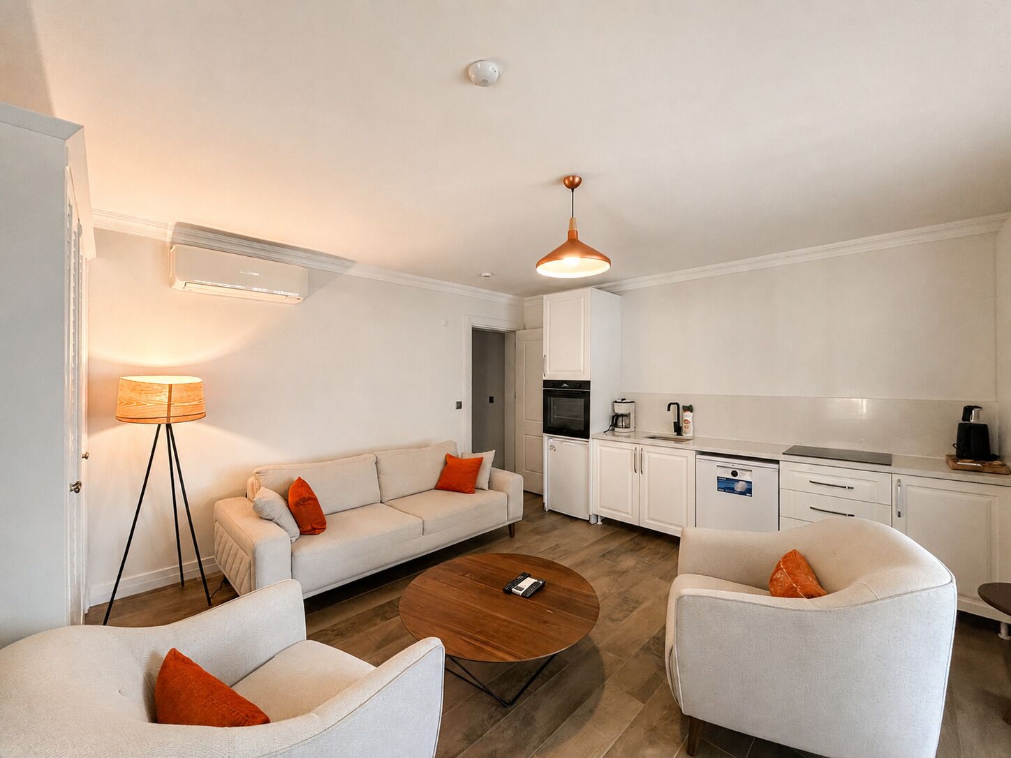 Una sala de estar moderna con un sofá, sillones, una mesa de centro y una zona de cocina con armarios y electrodomésticos.