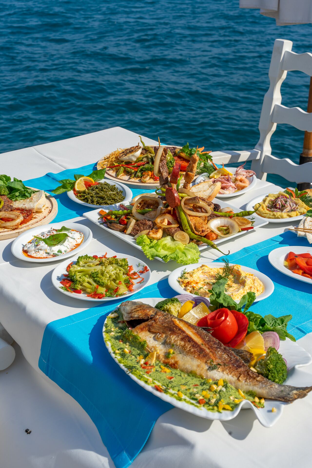 A table set with various dishes, including grilled fish, salads, and skewers, on a boat with a view of the sea.