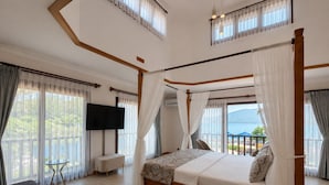 Una cama con dosel y cuatro columnas, un televisor de pantalla plana, una pequeña mesa redonda y un sofá en una habitación con ventanas amplias y un balcón.