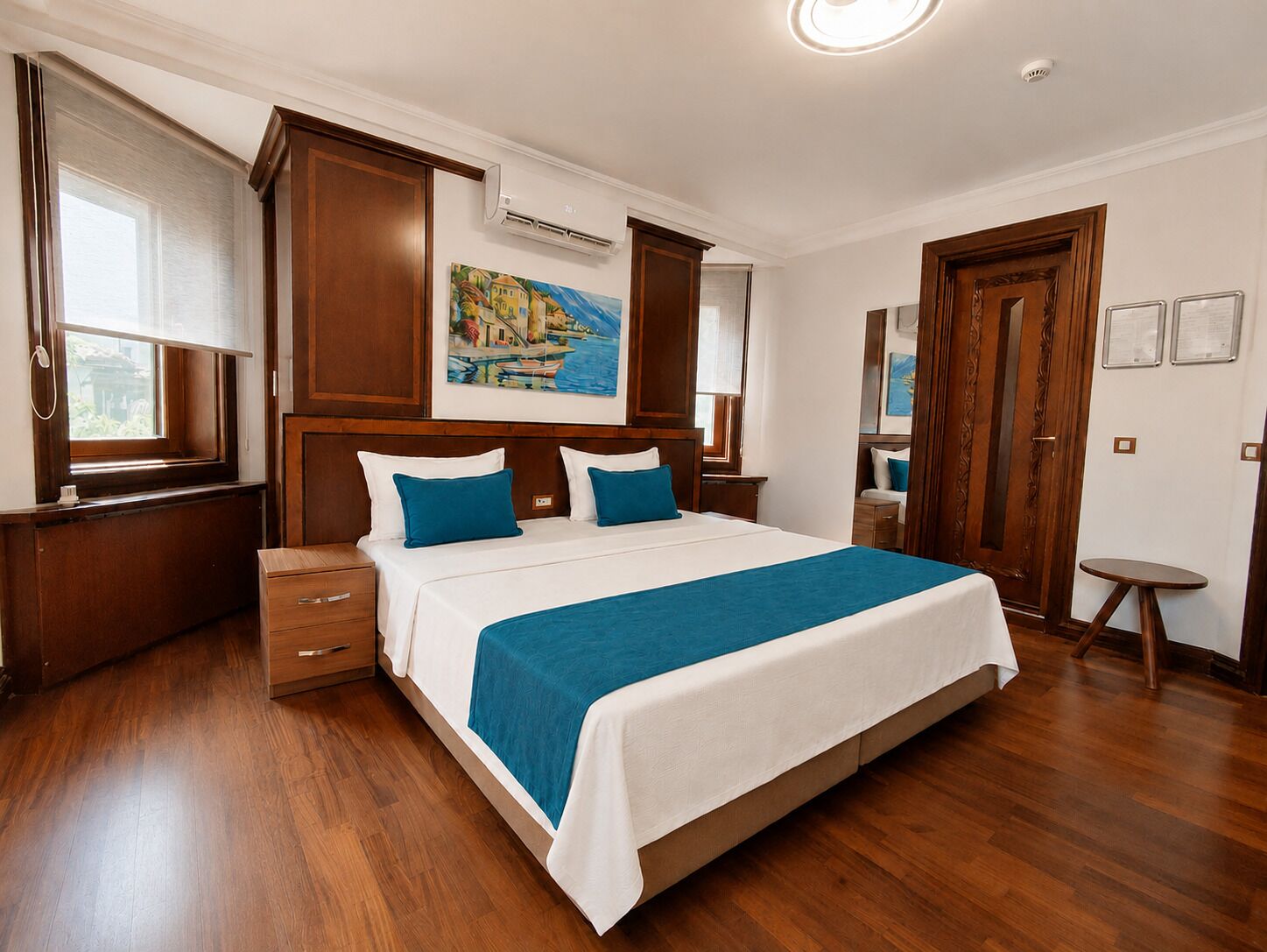 A hotel room with a large bed, wooden floor, a painting on the wall, and a window with blinds.