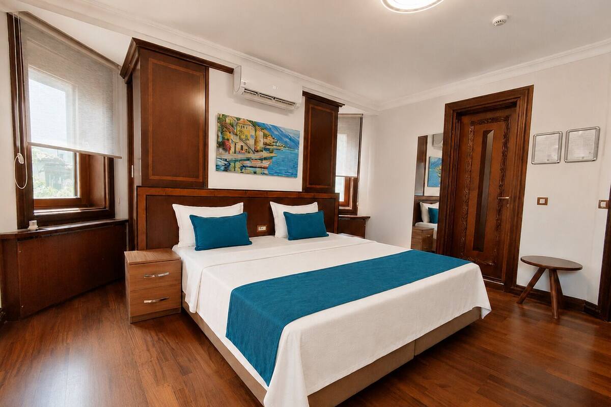 Habitación de hotel con una cama grande, suelo de madera, un cuadro en la pared y una ventana con persianas.
