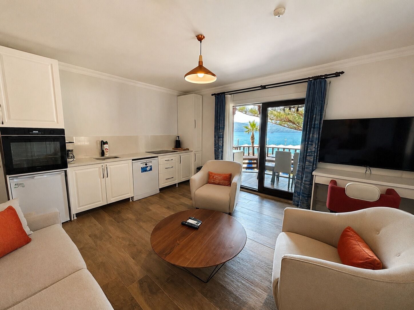 A modern kitchen with white cabinets, a black oven, and a white dishwasher. There is a round wooden coffee table in the center of the room, flanked by two beige sofas with orange cushions. A large flat-screen TV is mounted on the wall to the right.