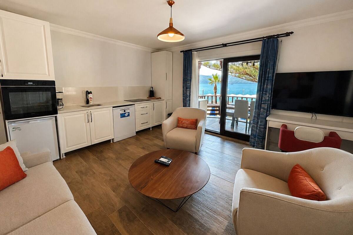 Una cocina moderna con armarios blancos, un horno negro y un lavavajillas blanco. En el centro de la habitación, hay una mesa de centro redonda de madera, flanqueada por dos sofás beige con cojines naranjas. Un televisor de pantalla plana grande está montado en la pared a la derecha.