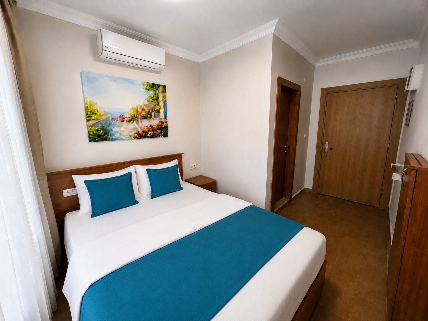 A hotel room with a bed, a painting, an air conditioning unit, a wardrobe, and a door.