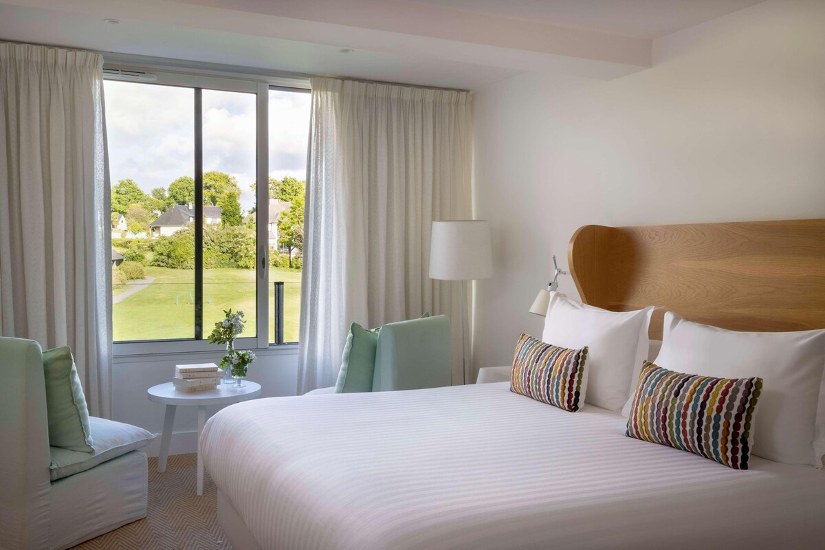 A hotel room with a large bed, a small table, a chair, and a view of a golf course.