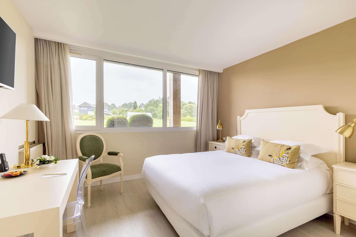 Habitación de hotel con una cama grande, un escritorio con lámpara, una silla y vistas a zonas verdes.