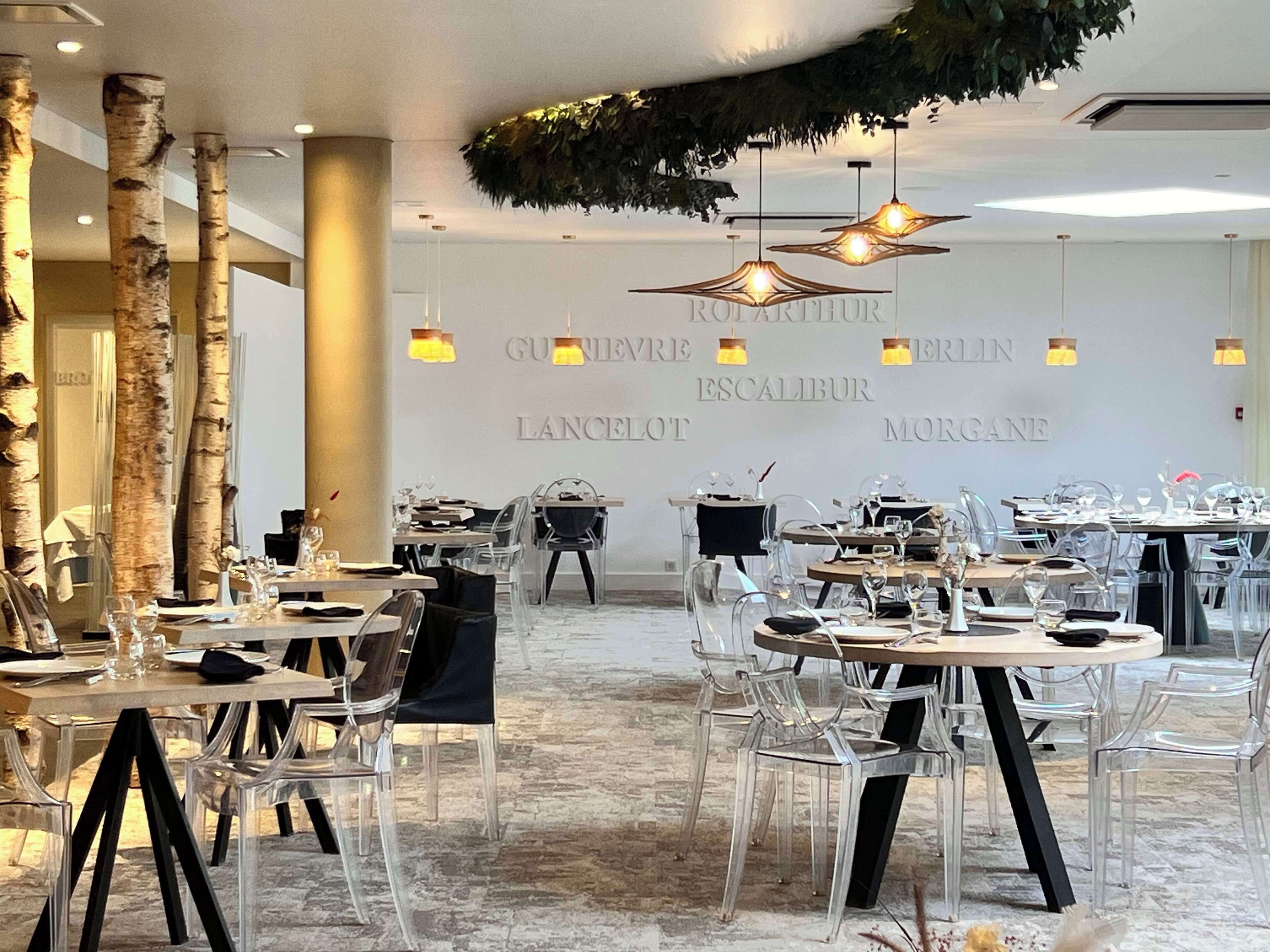 A restaurant with clear glass tables and chairs, a stone floor, and a wall with the name 'GUESTHOUSE' and 'ESCALABUR' visible.