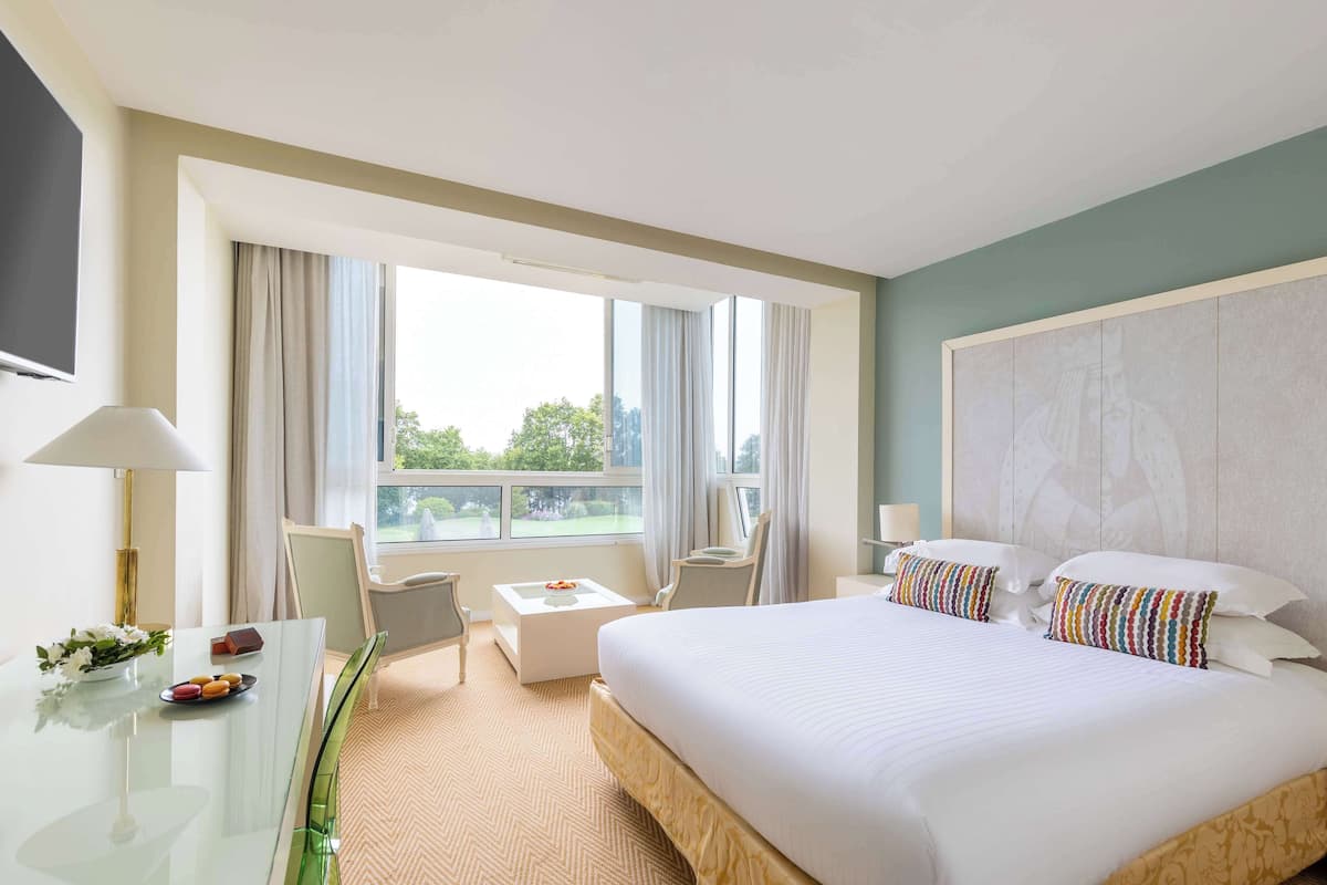 Habitación de hotel con una cama grande, un escritorio con lámpara, una silla, una mesita con un florero y vistas al exterior a través de amplios ventanales.