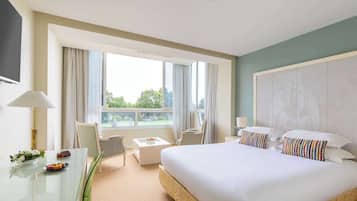 A hotel room with a large bed, a desk with a lamp, a chair, a small table with a flower vase, and a view of the outdoors through large windows.
