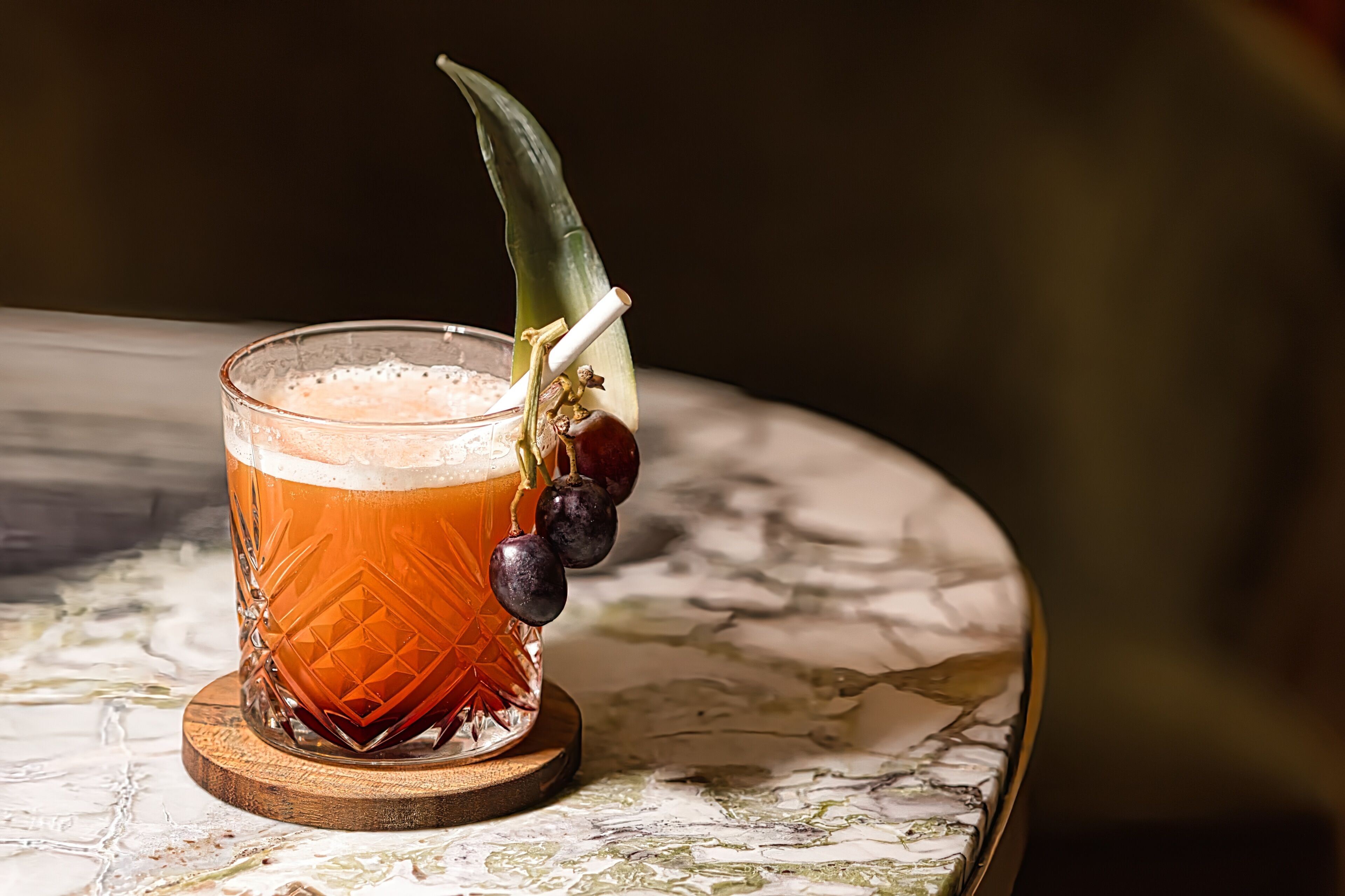 A glass of amber-colored beverage with a decorative leaf and grapes on a coaster.