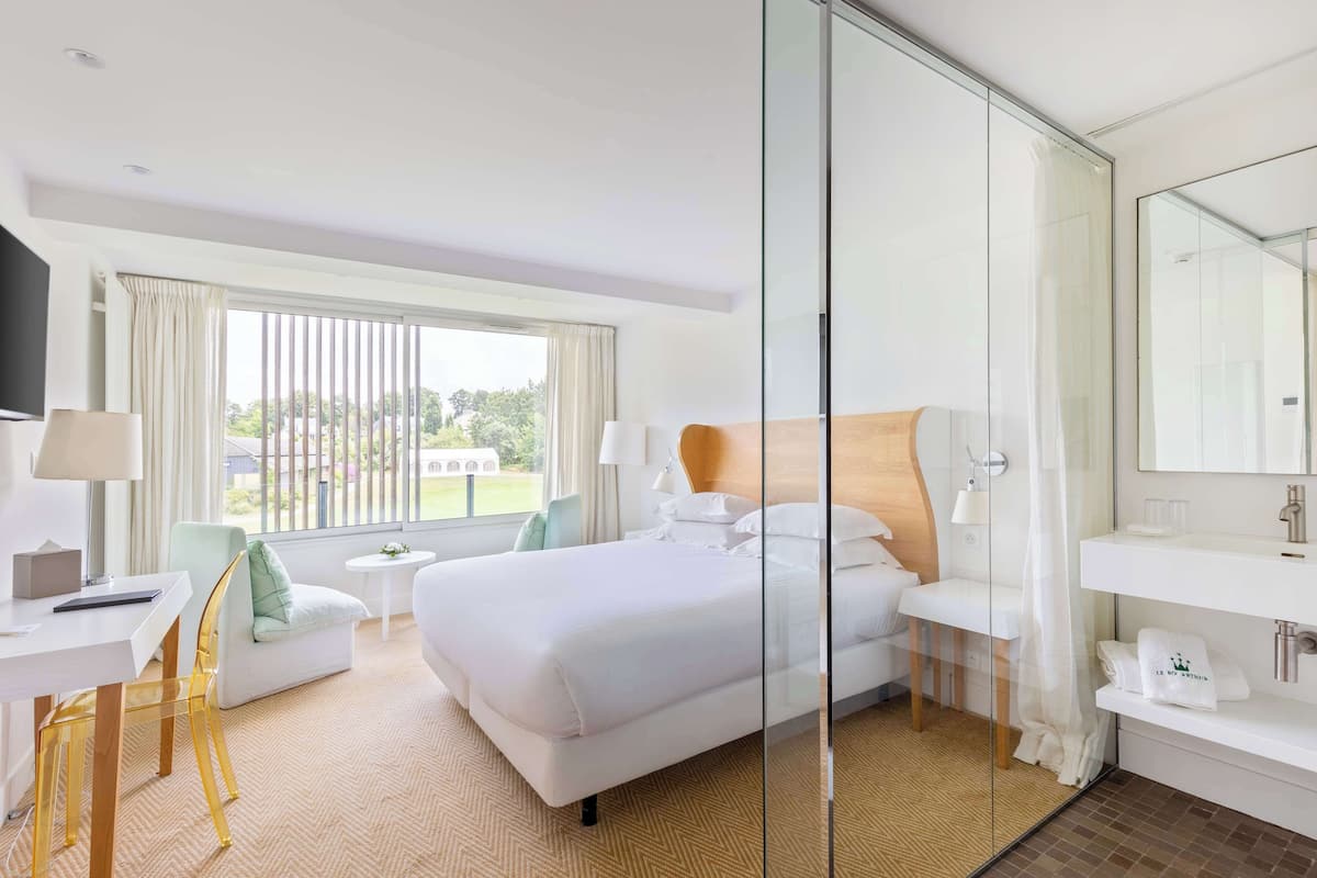 Una habitación de hotel moderna con una cama grande, una zona de estar con una silla y una mesa, un separador de vidrio y un baño con espejo y lavamanos.