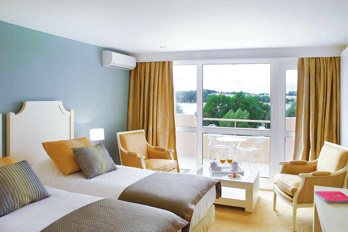 Habitación de hotel con dos camas, un área de estar con una mesa y sillas, y un ventanal con cortinas.