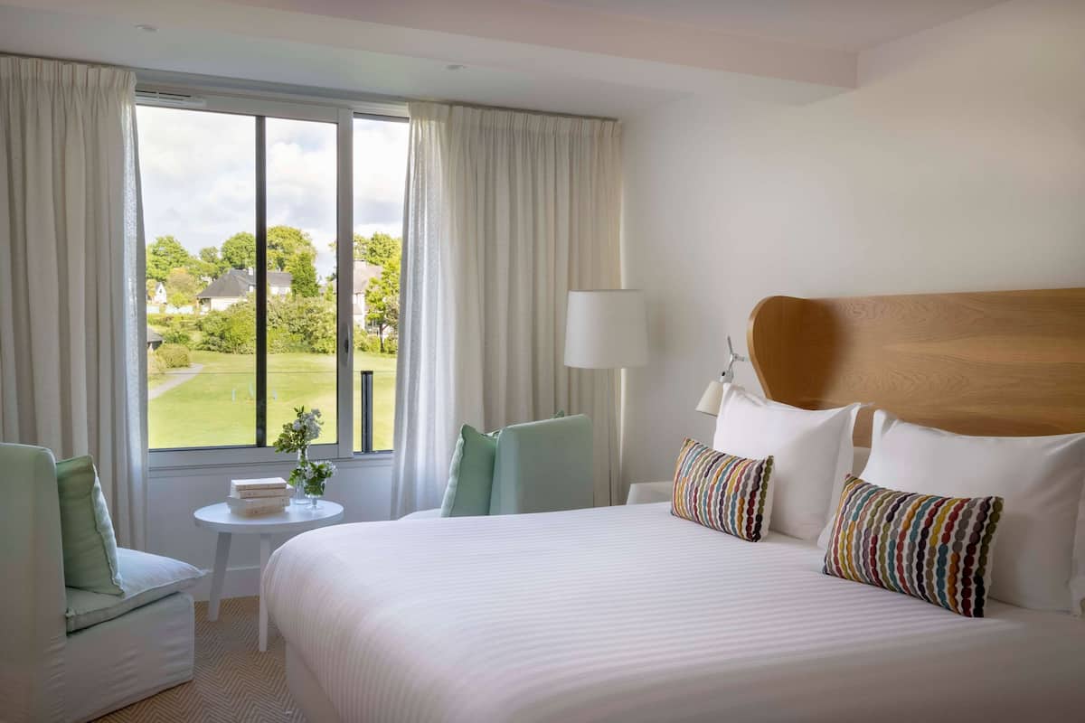 Habitación de hotel con una cama grande, una mesita con un jarrón, una silla y vista a un paisaje verde a través de la ventana.