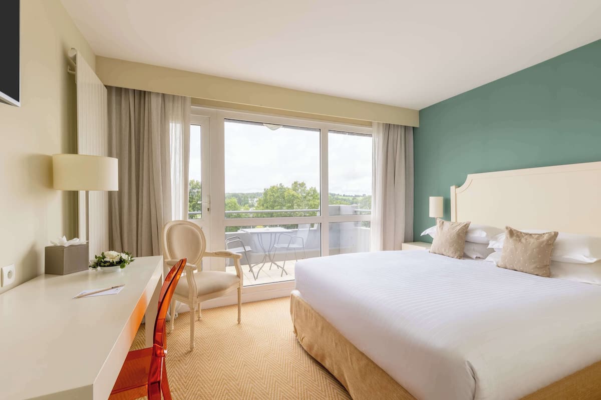 Habitación de hotel con una cama grande, un escritorio con silla, un balcón con vistas y una ventana con cortinas.