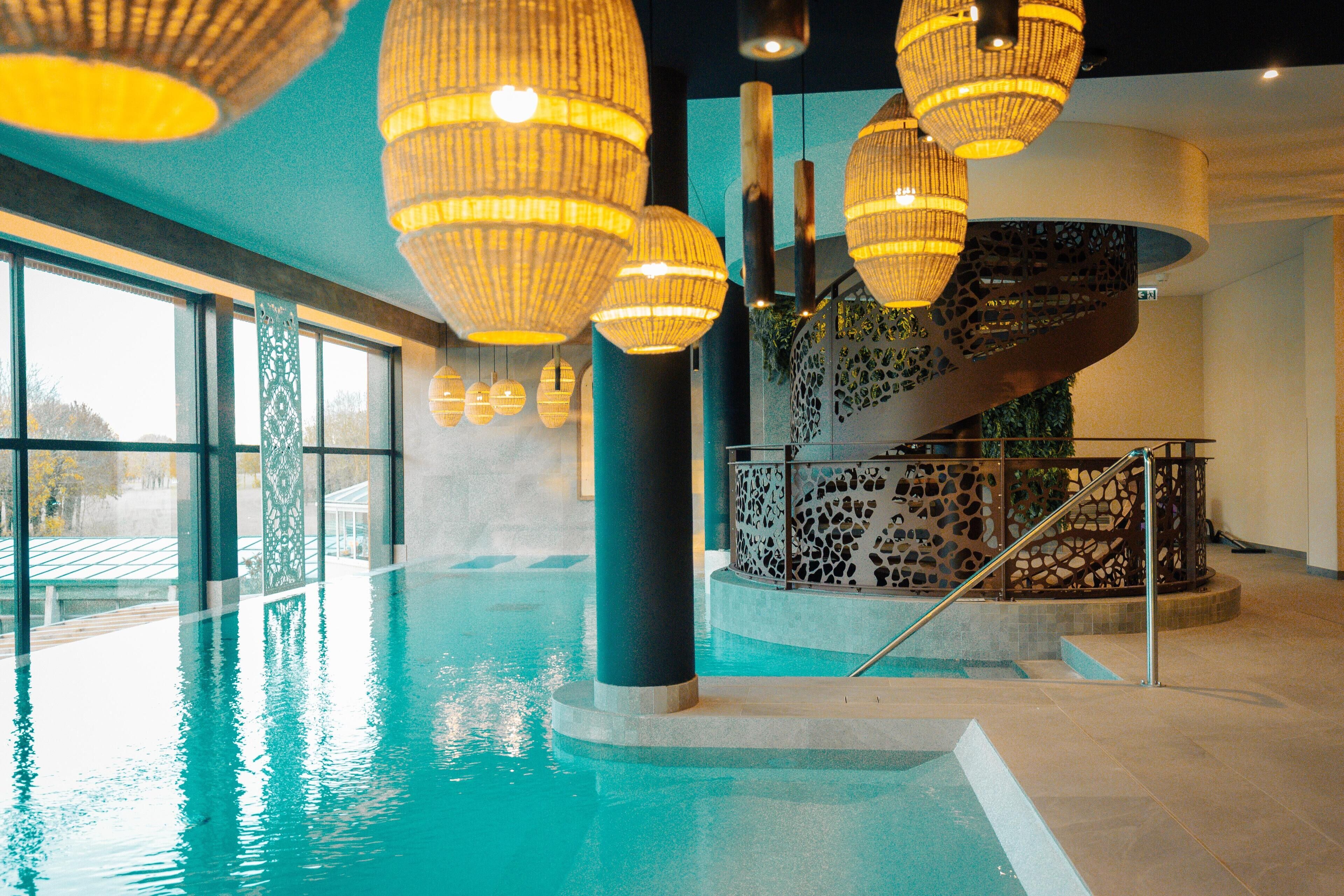 Una piscina interior con paredes de vidrio transparente, una escalera con barandilla decorativa y varias lámparas colgantes.
