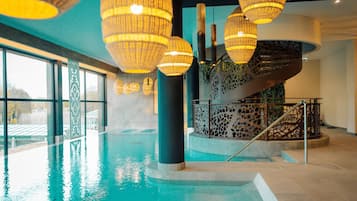 Una piscina interior con paredes de vidrio transparente, una escalera con barandilla decorativa y varias lámparas colgantes.