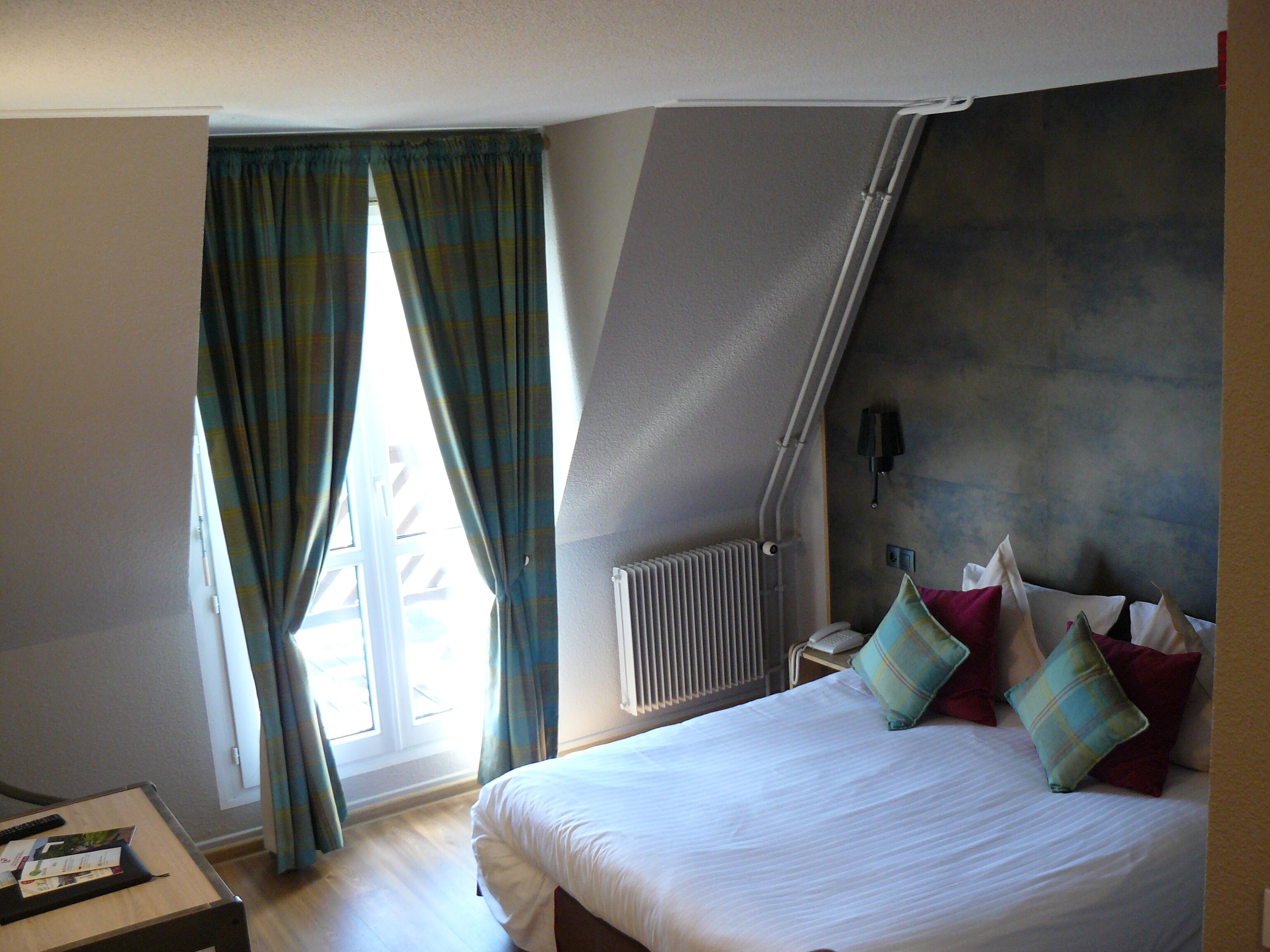 Une chambre d’hôtel comprenant un lit, un bureau, une fenêtre avec des rideaux, un radiateur et une lampe fixée au mur.
