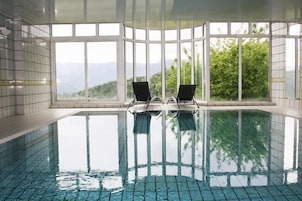 Una amplia piscina interior con paredes de vidrio transparente, dos sillones de descanso y vistas a zonas verdes y montañas.