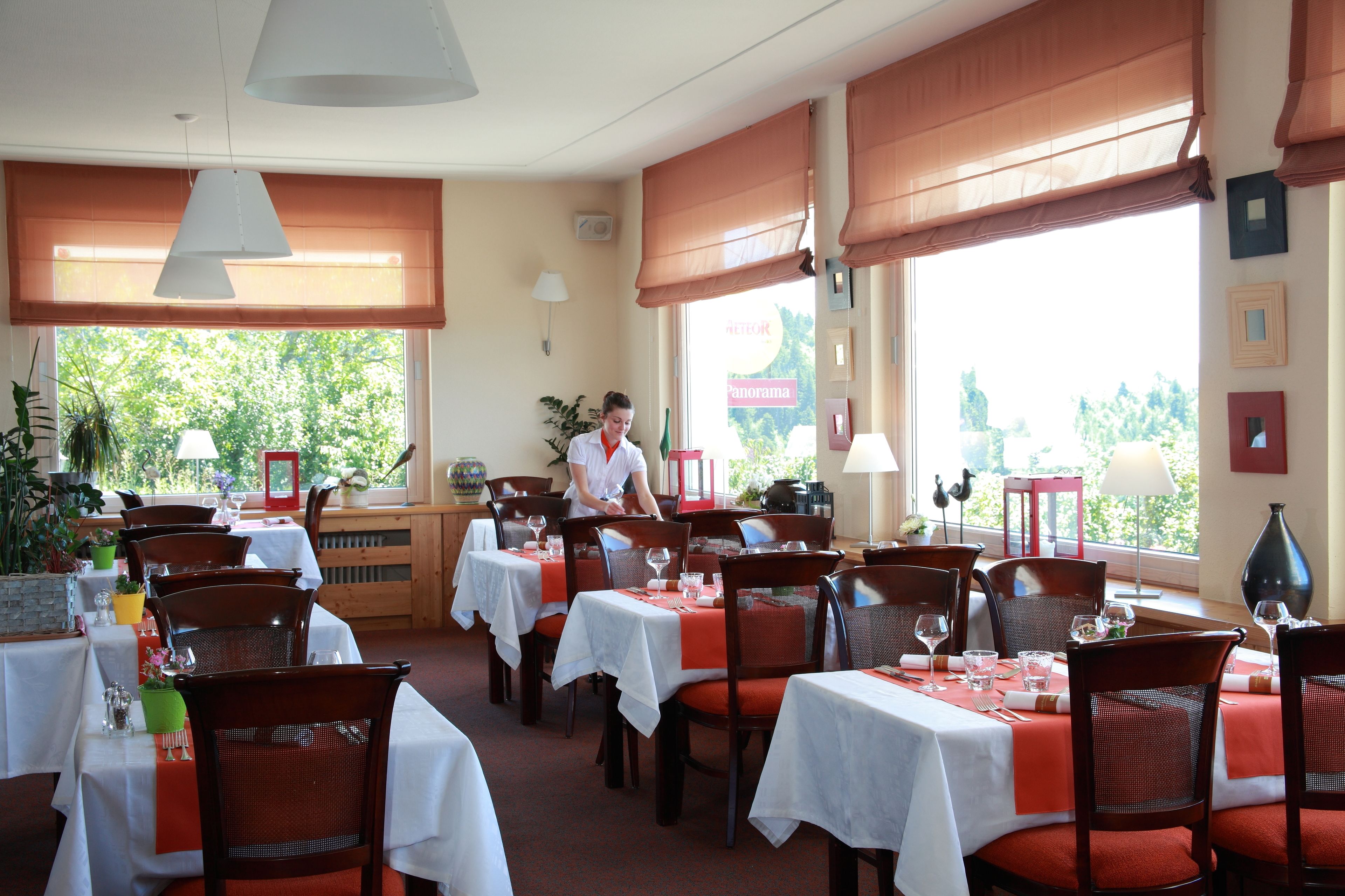 Een restaurant met gedekte tafels, een ober die gasten bedient, en grote ramen met uitzicht op groen.