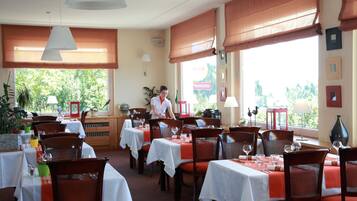 Een restaurant met gedekte tafels, een ober die gasten bedient, en grote ramen met uitzicht op groen.