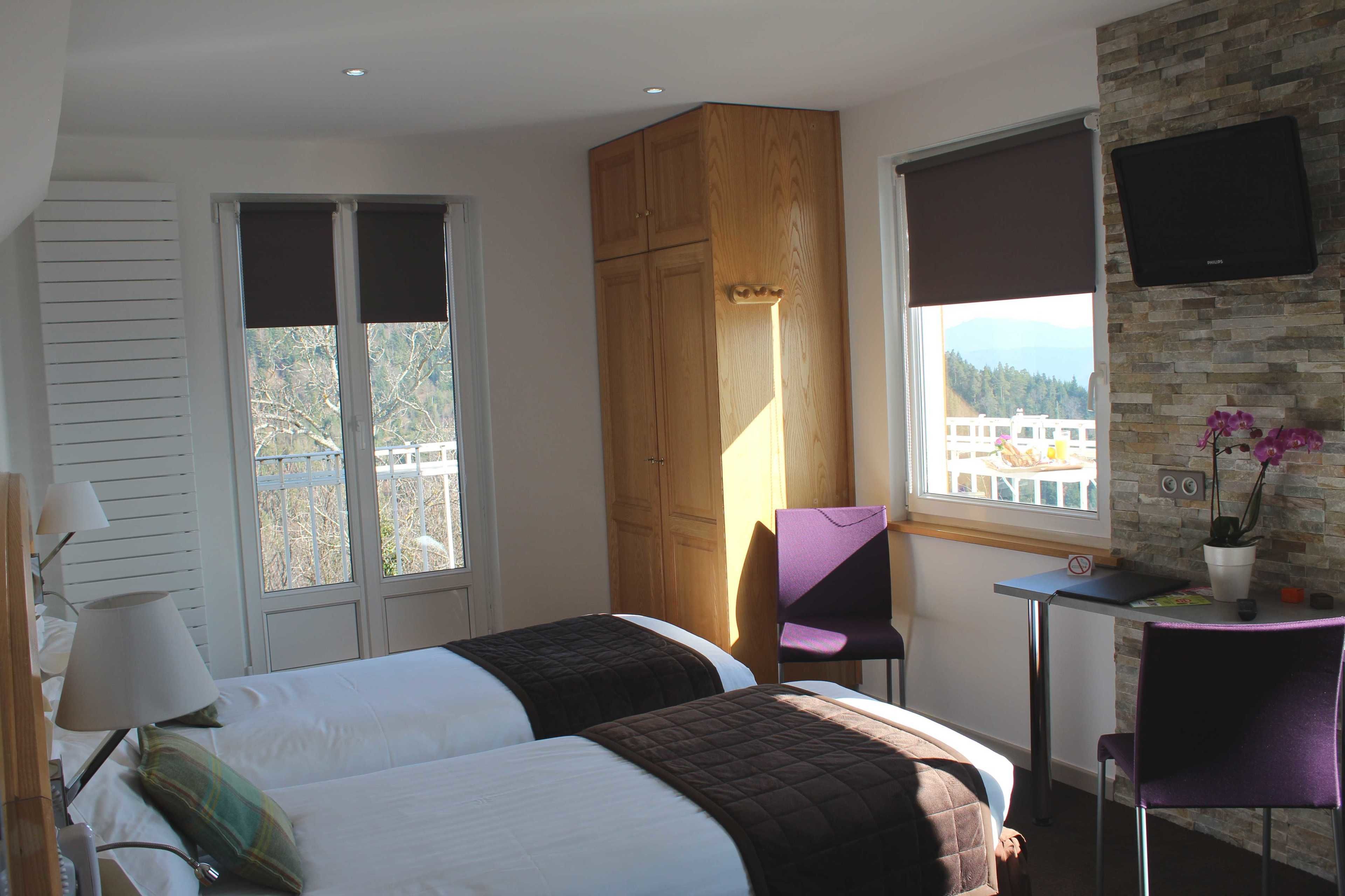A hotel room with two beds, a wooden wardrobe, a desk with a chair, a TV, and a window with a view of a building and trees.