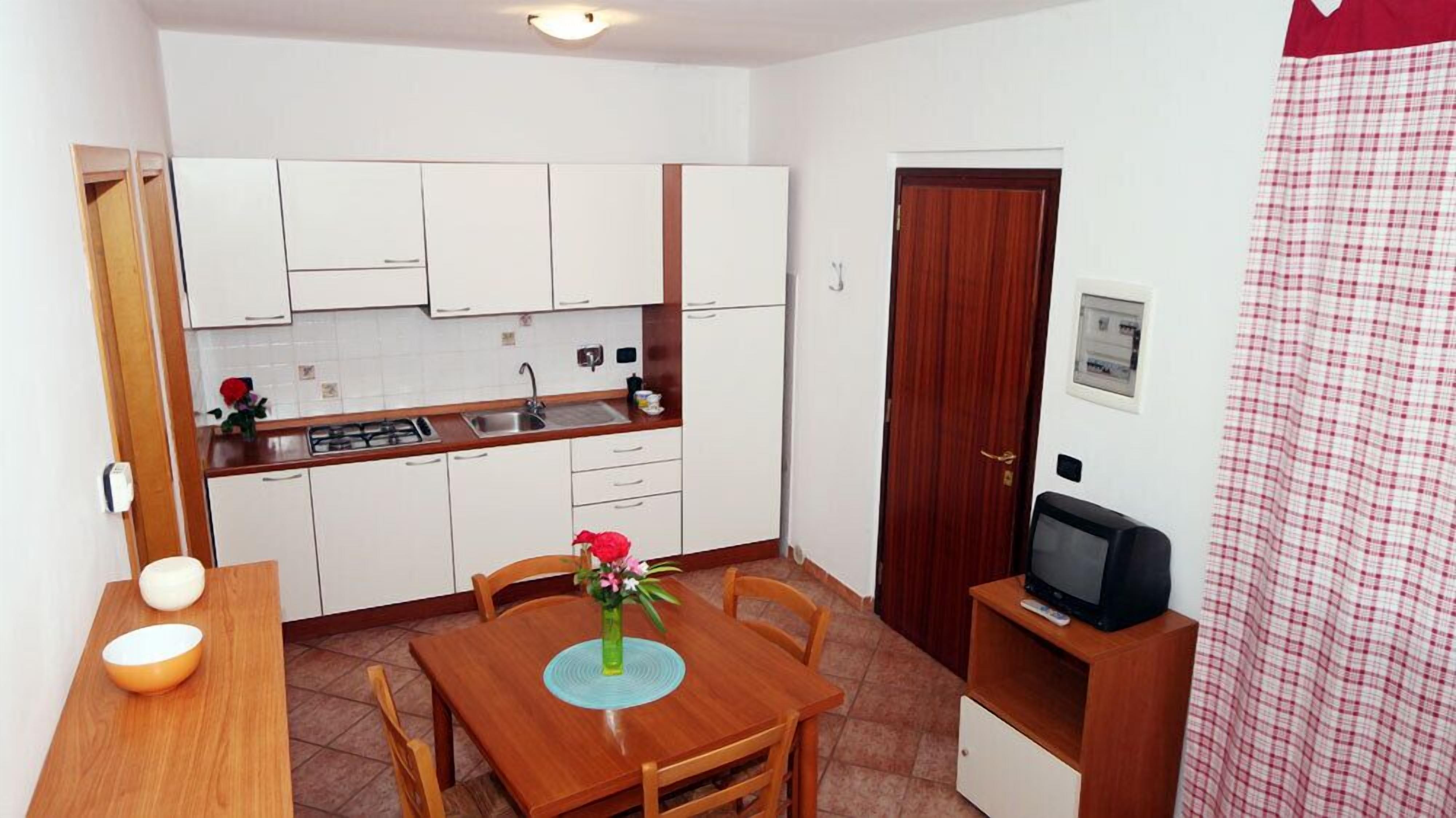 Een kleine keuken met witte kastjes, een houten eettafel met stoelen, een kleine televisie op een houten dressoir, en een rood-wit geruit gordijn.