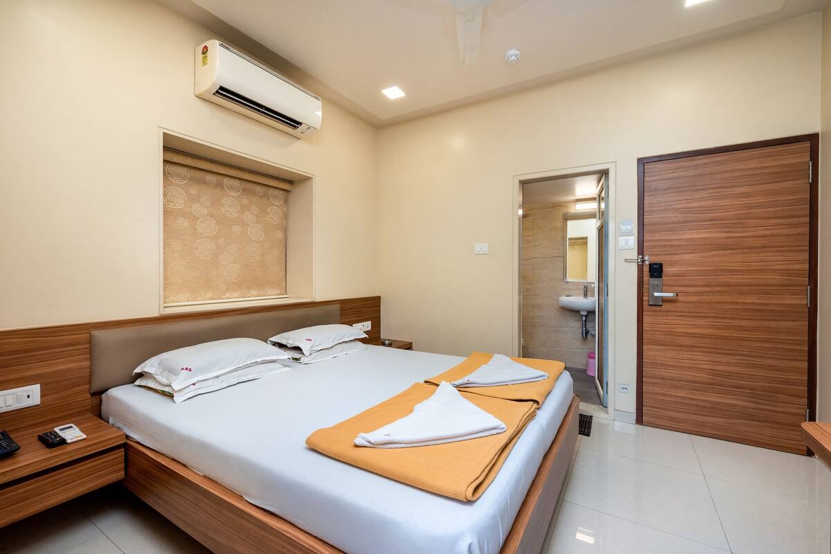 Une chambre d’hôtel avec un lit, des tables de chevet, un climatiseur, une porte en bois et une salle de bain visible à travers une porte ouverte.