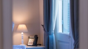 A hotel room with a bedside table, a lamp, a telephone, and a window with curtains. - Palladian Home (Athens)