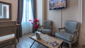 A hotel room with a wooden bench, two armchairs, a coffee table with a wine bottle and glasses, a TV on the wall, and a window with curtains.