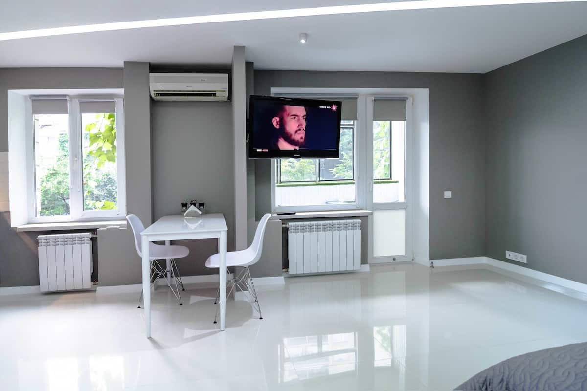 A modern room with a flat-screen TV mounted on the wall, a white table with two chairs, and large windows providing natural light.