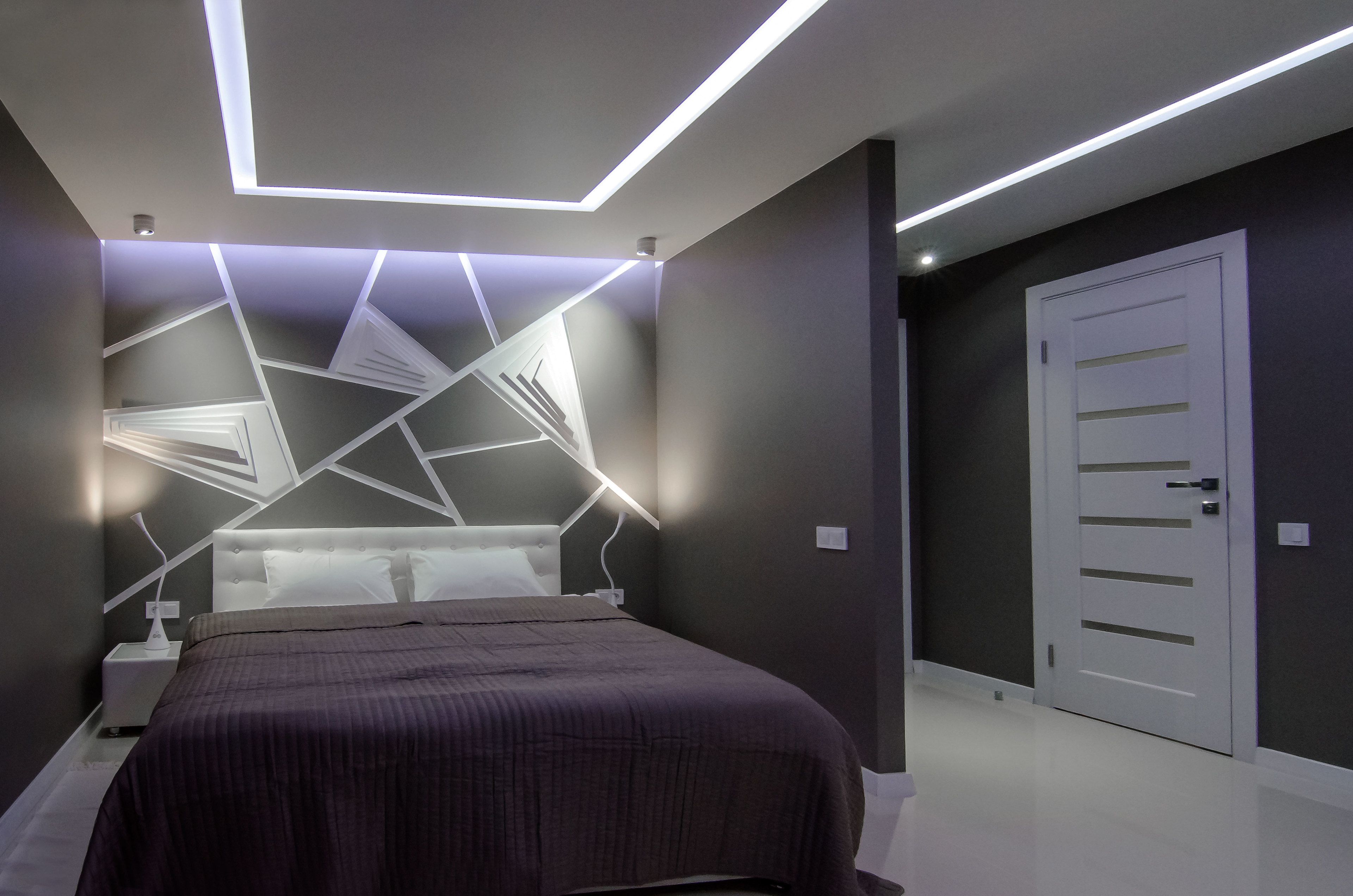 Een moderne slaapkamer met een geometrisch plafondontwerp, een bed met een donkerdekkentje en een witte deur.