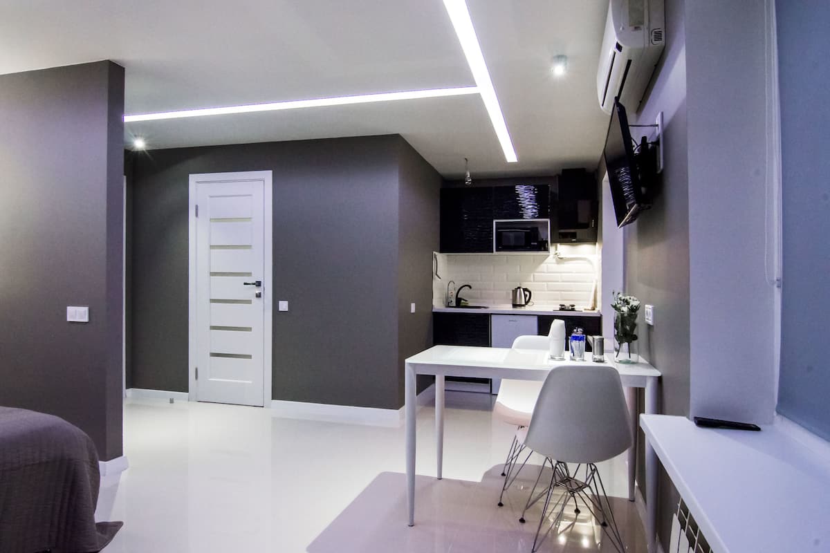 A modern, well-lit room with a white desk, grey chairs, a white door, and a kitchen area with a microwave and sink.